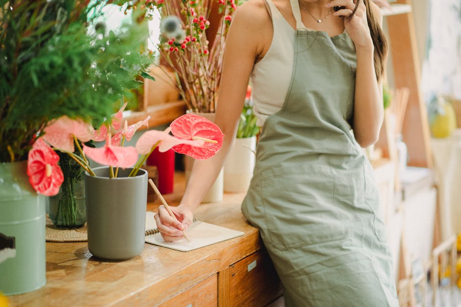 pexels-photo-5410105-5410105 Crop faceless female florist in apron standing near wooden table with flowers and writing notes in notebook while talking on mobile phone